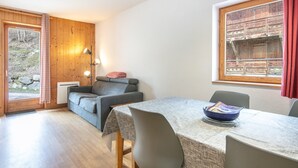 A cozy room with a sofa, a dining table with chairs, a window with a view of a wooden structure, and a door leading outside. - Apartment in Saint-sorlin Near Ski Slopes (Saint-Sorlin-d'Arves)