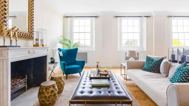 A spacious living room with a fireplace, a blue armchair, a sofa with patterned cushions, and a large mirror.