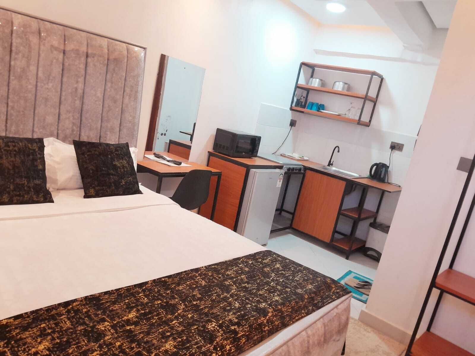 Une chambre d’hôtel de taille réduite, comprenant un lit, un bureau, une kitchenette et un petit réfrigérateur.