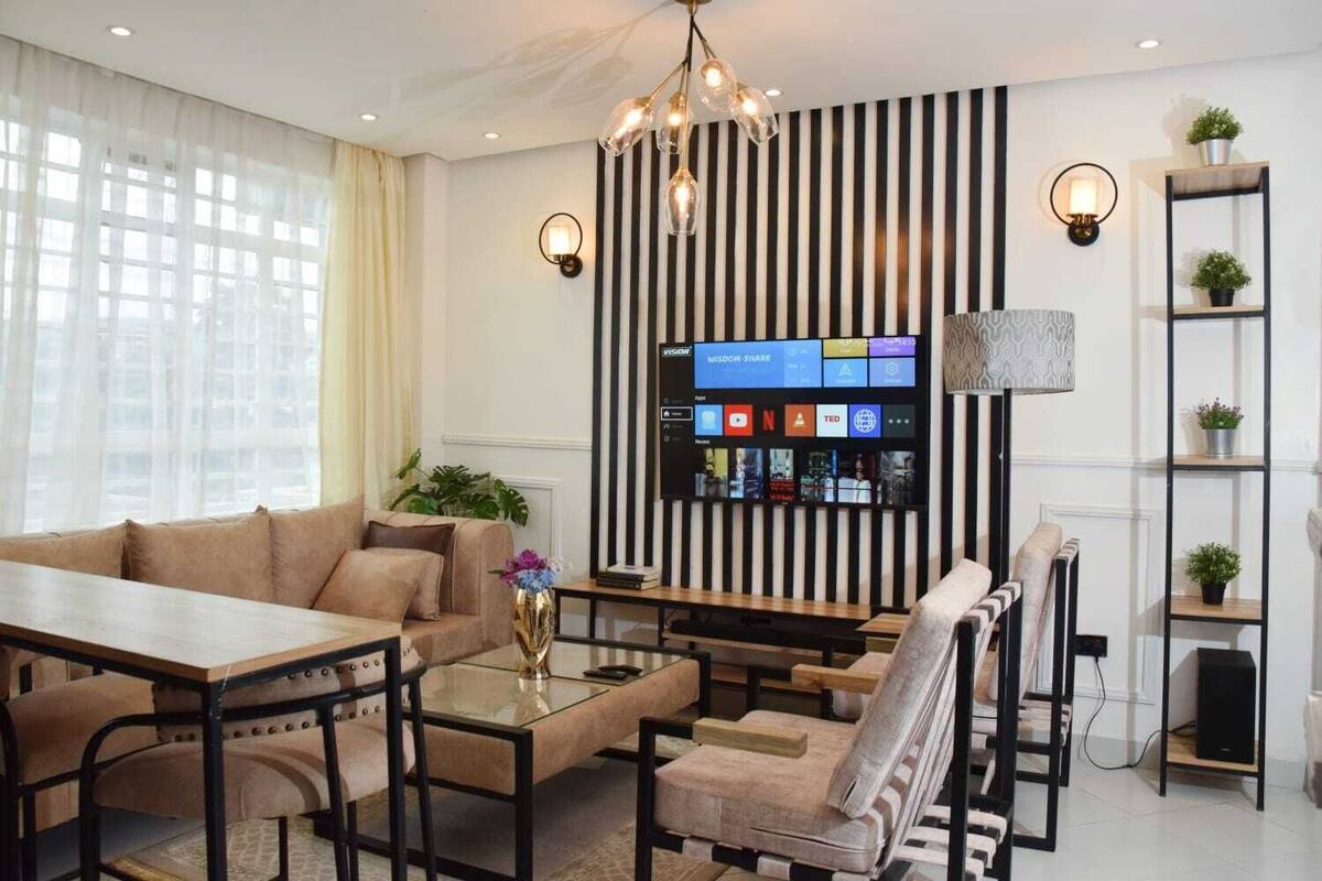 Una sala de estar moderna con sofá, mesa de centro y sillas. Un televisor está montado en la pared mostrando varios canales.