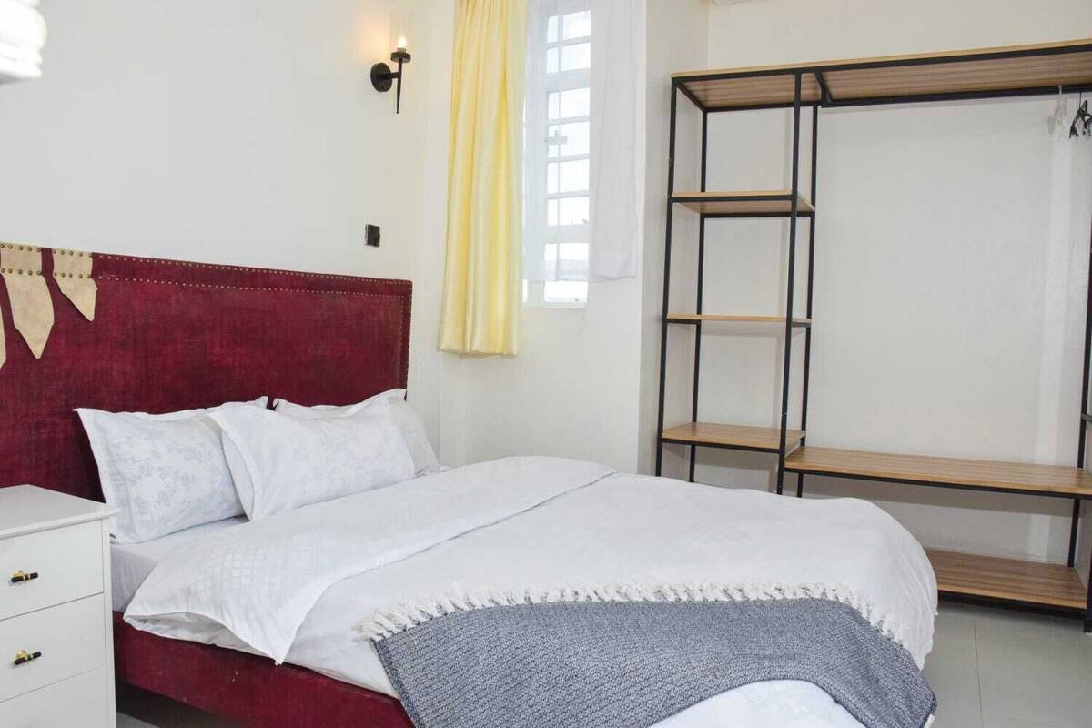 Un dormitorio con cabecera roja, ropa de cama blanca, un plaid gris, una estantería de madera y una mesita de noche blanca con cajones.