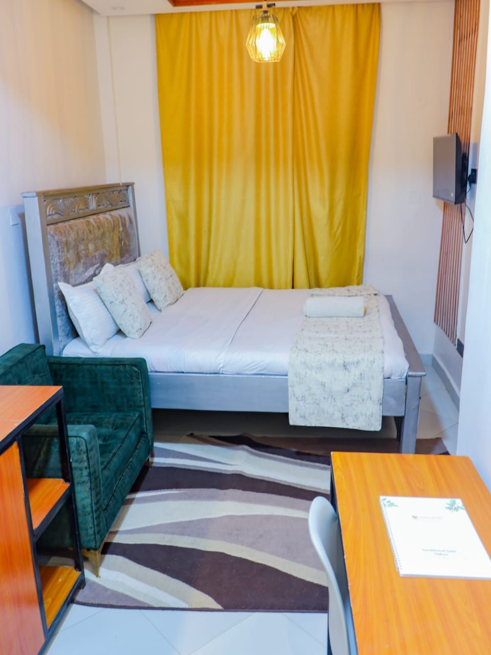 A small, compact room with a bed, a green armchair, a wooden table, and a patterned rug.