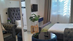 A modern hotel room with a large bed, a sofa, a chair, and a small table. There is a TV mounted on the wall and a window with curtains. - Sandalwood Suites Nakuru (Nakuru)