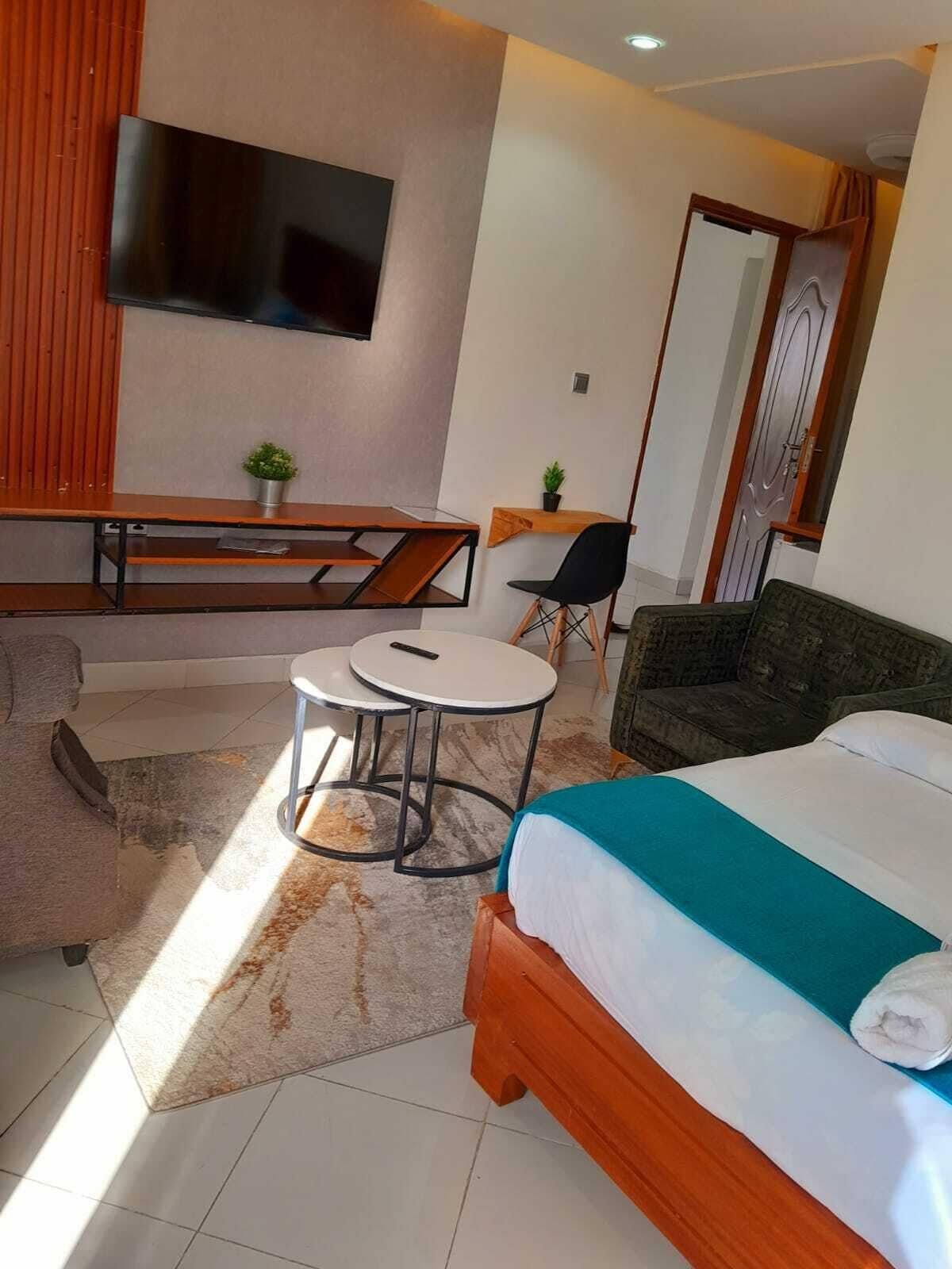 A modern hotel room with a bed, a sofa, a small round table, a TV mounted on the wall, and a wooden bench.