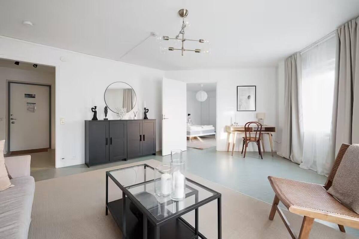A modern hotel room with a glass coffee table, a sofa, a dining area with a table and chairs, a wardrobe, and a large mirror.
