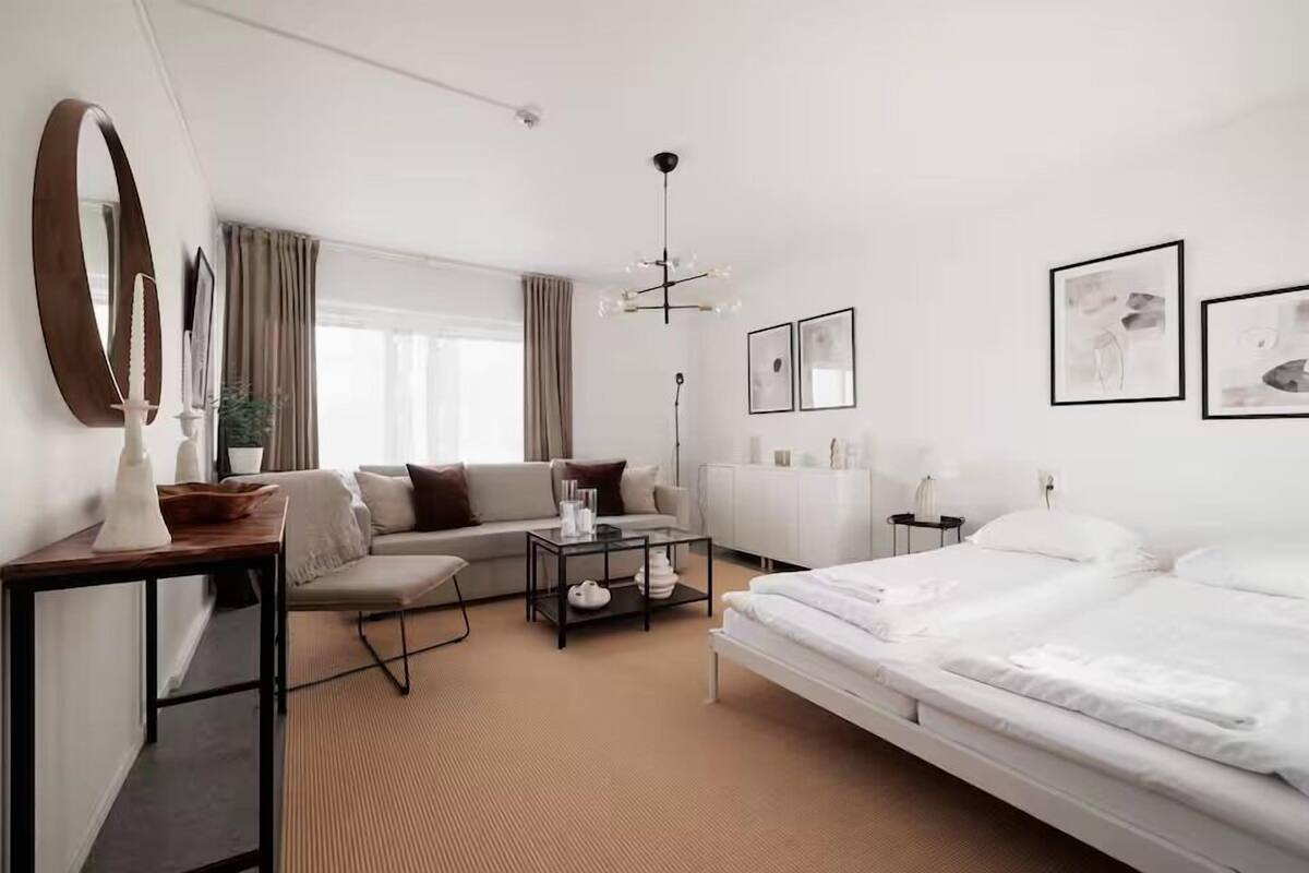 A modern hotel room with a large bed, a sofa, a small table, and a desk. There are framed pictures on the wall and a large mirror on the wall.