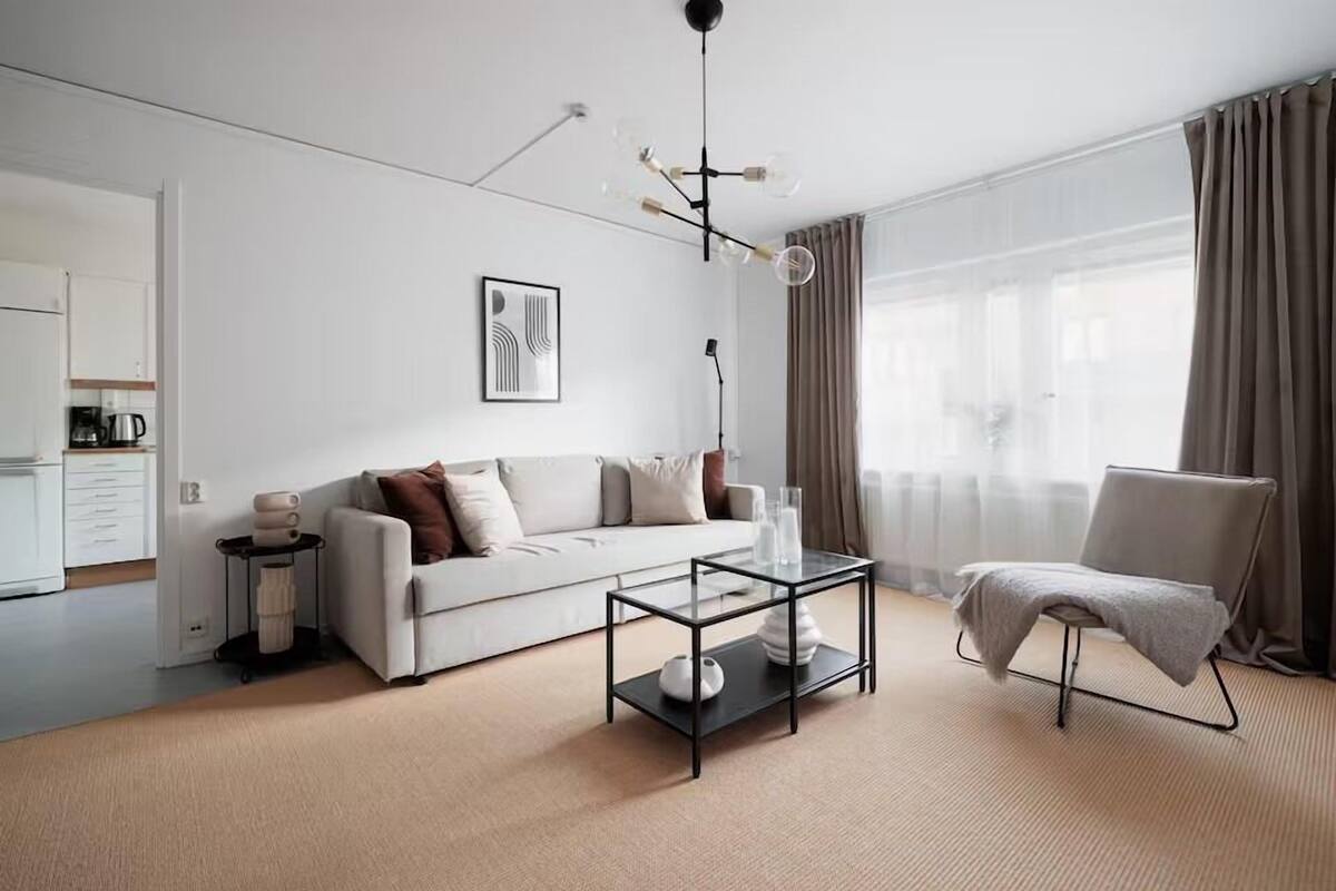 A modern living room with a sofa, a glass coffee table, a chair, and a dining area visible through an open door.