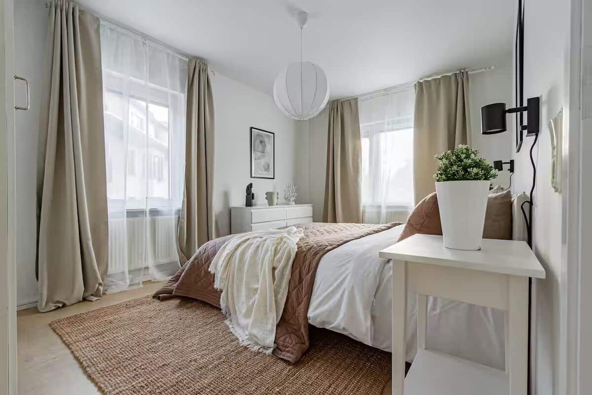 A bedroom with a bed, bedside table, lamp, curtains, and a window.