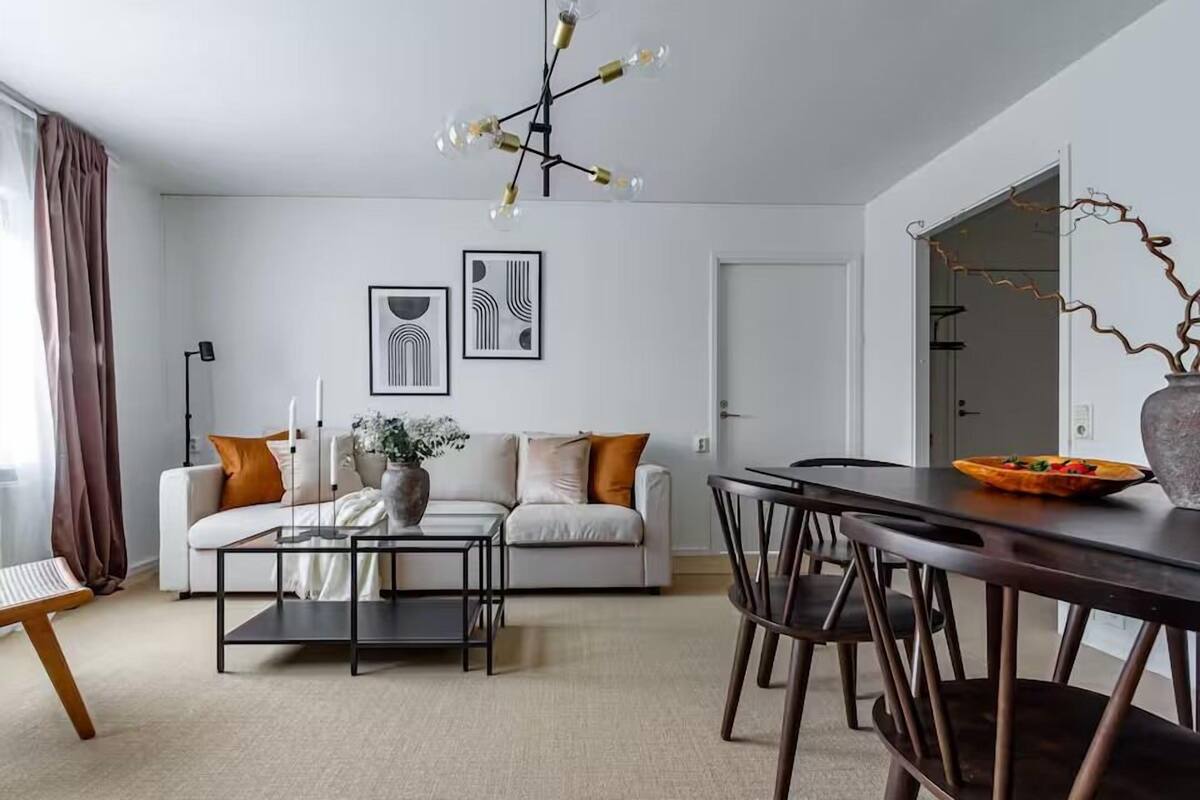 A modern living room with a sofa, coffee table, and dining table. There are framed artworks on the wall and a vase with branches on the dining table.