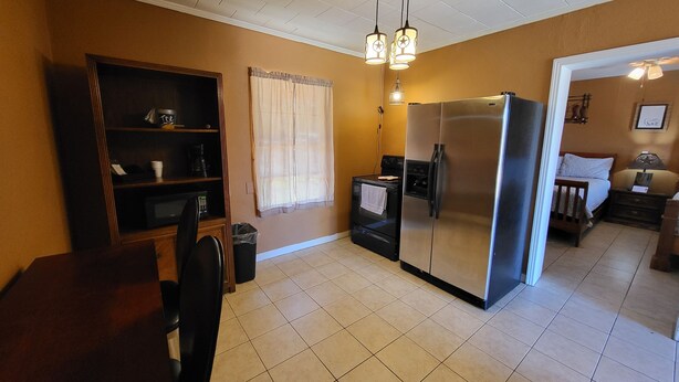 A compact kitchen with a refrigerator, stove, and microwave. There is a wooden cabinet and a trash can. The room has a tiled floor and a window with curtains.