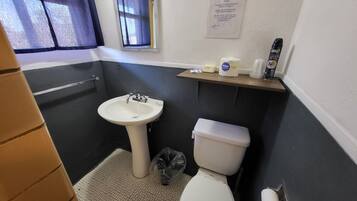 Un baño con un lavamanos blanco, un inodoro y una pequeña ventana con cortina. Hay un aviso en la pared y un estante con artículos de aseo.