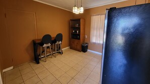 A room with a tiled floor, a dining table with chairs, a cabinet, a trash bin, and a refrigerator.