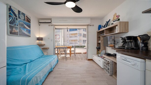 A compact living space with a blue sofa, kitchenette, and a balcony with a view of a building. - Apartment in Agde Near Beach & Heated Pool (Agde)
