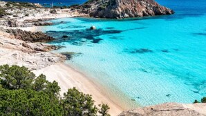 A coastal landscape with a sandy beach, clear turquoise waters, and rocky terrain. - Cozy Flat Just Steps From Baia Delle Mimose (Badesi)