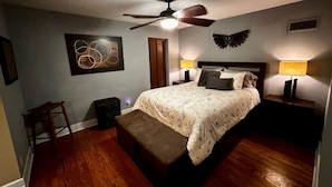 A bedroom with a bed, two bedside tables with lamps, a ceiling fan, a wooden floor, a small stool, and a wall art piece.