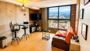 A compact living space with a kitchenette, a large window with a city view, a leather sofa, and a TV mounted on the wall.