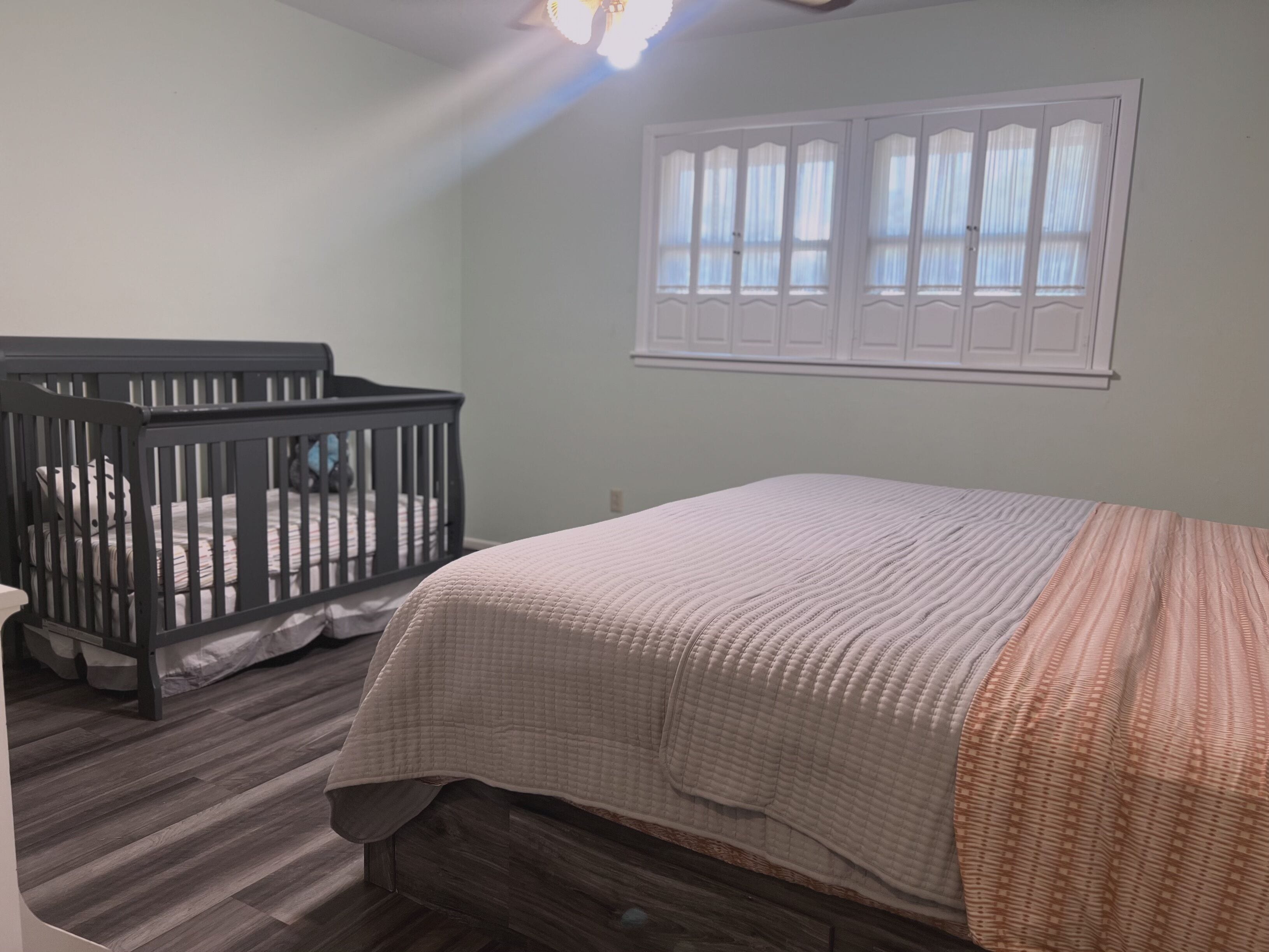 A bedroom with a crib, a bed with a quilt, wooden flooring, and a window with curtains.