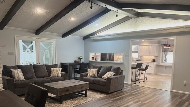 A spacious living area with a dark wood floor, a large gray sectional sofa, a wooden coffee table, and a dining table with chairs. There is a kitchen area visible through an open doorway. - Reserved Luxury (Lubbock)
