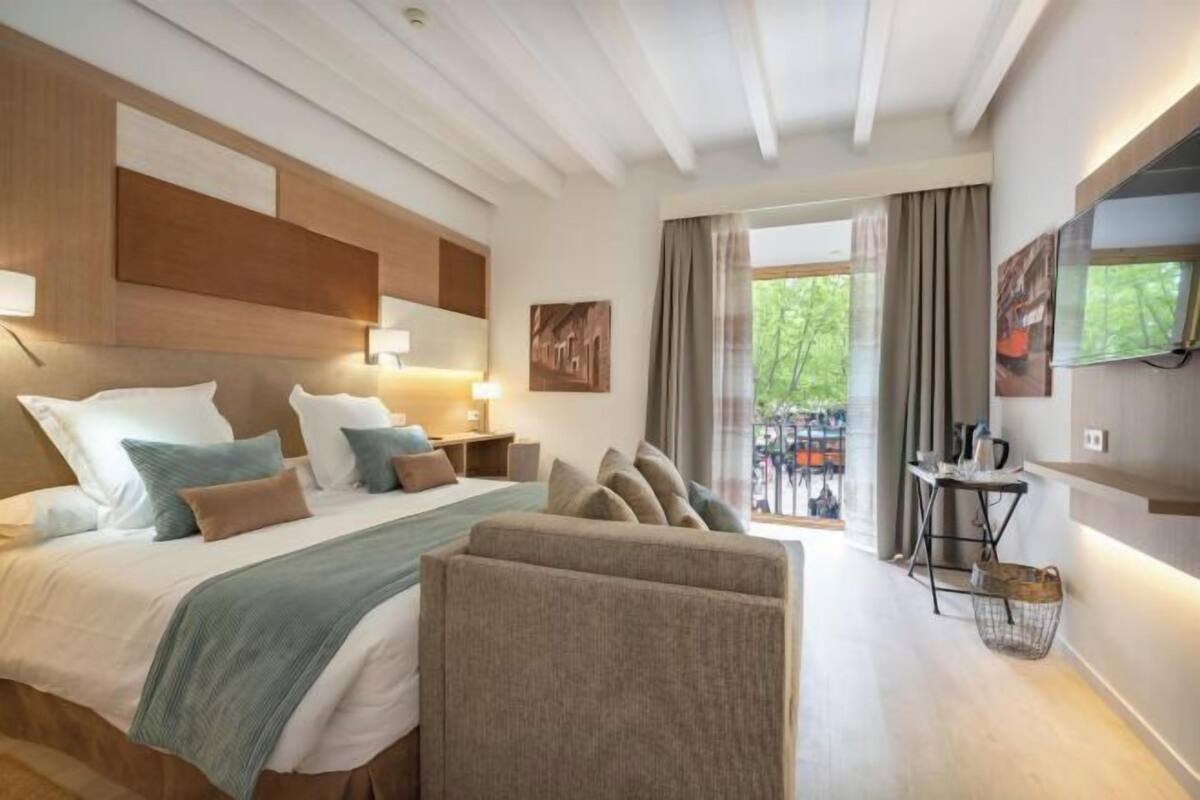 A modern hotel room with a large bed, a sofa, a desk, and a view of the outdoors.