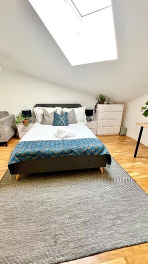 A bedroom with a skylight, a bed with a patterned bedspread, a grey rug, a grey armchair, a white dresser, and a small table with a plant. - Luxe Prima - 2 Bed in Central London (London)