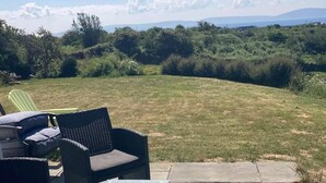 Un jardin avec une pelouse verdoyante, un espace patio aménagé avec du mobilier d’extérieur, et une vue sur un paysage naturel avec des arbres et des collines à l’horizon.