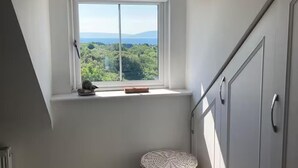 Une pièce dotée d’une grande fenêtre donnant sur une forêt et des montagnes, d’un radiateur blanc et d’un tabouret en osier.