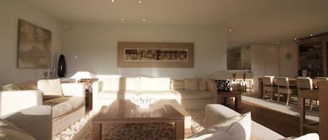A spacious living room with a large sofa set, a coffee table, and a dining area with chairs and a table.