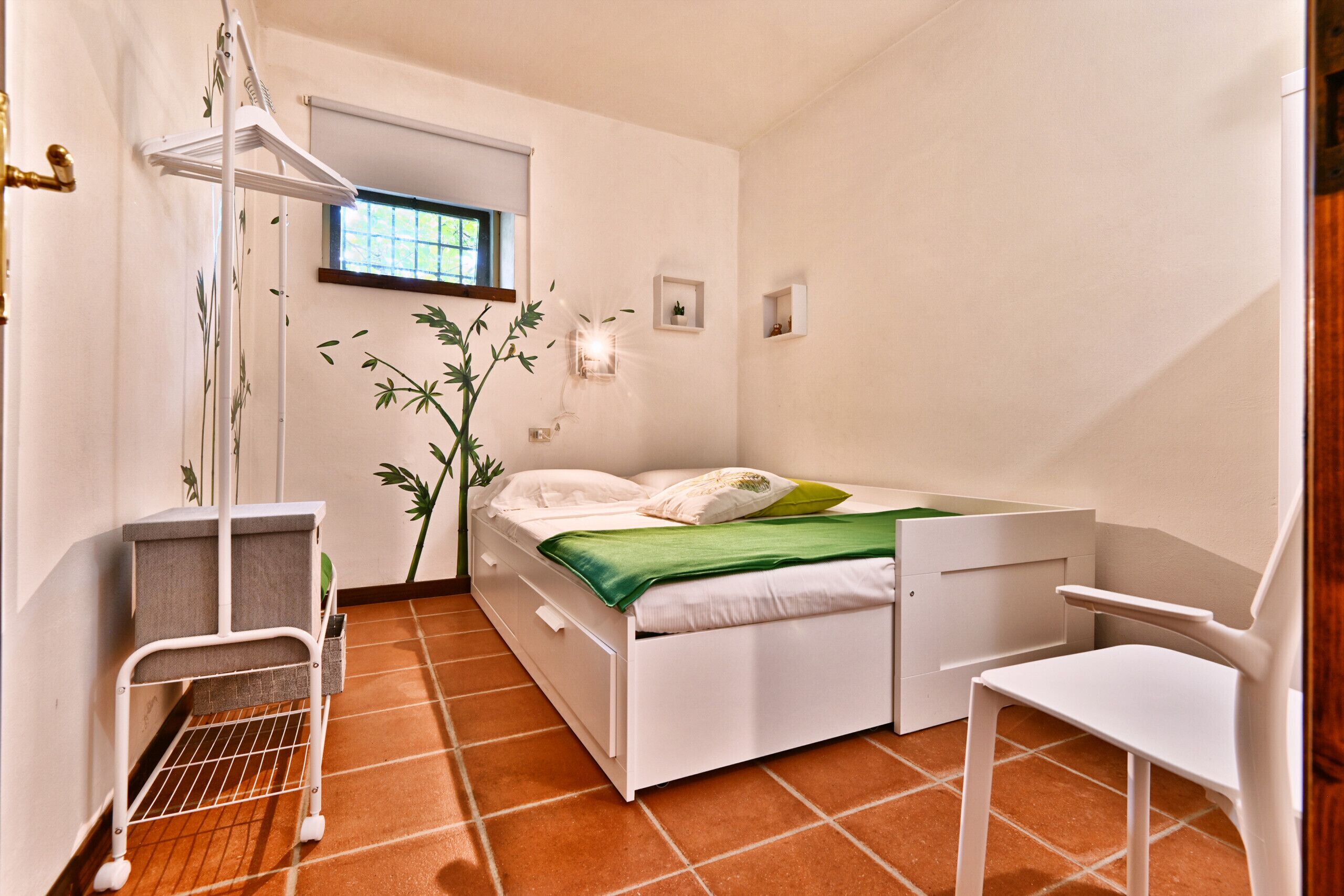 Una habitación pequeña y limpia con una cama, una silla, una mesita y un ventanal.
