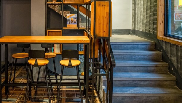 Un interior moderno con una escalera de madera, una zona de comedor con taburetes y una estantería.