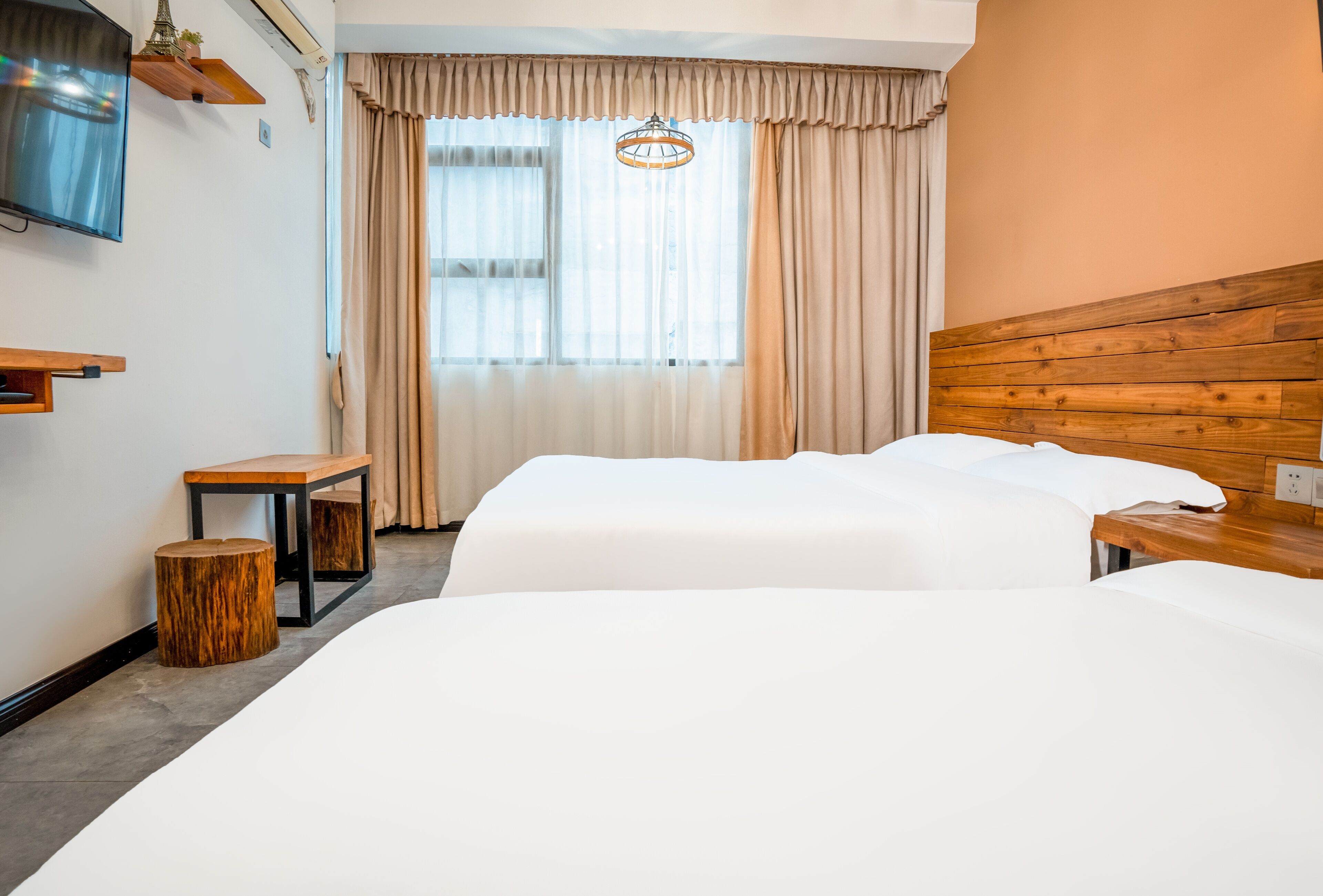 A hotel room with two beds, a TV, a wooden headboard, a bedside table, a small stool, and a window with curtains.