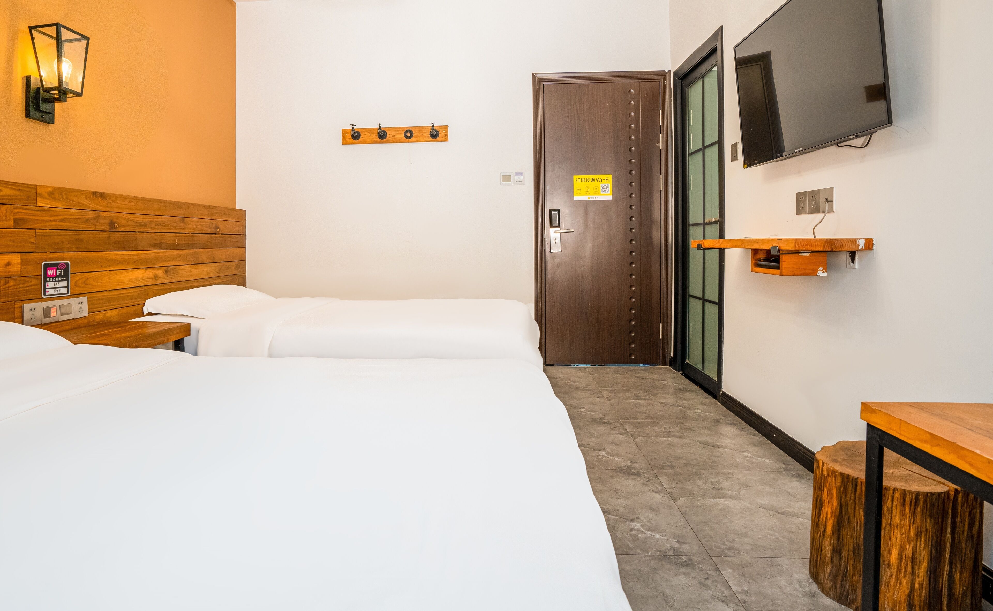 A hotel room with two beds, a wooden headboard, a TV mounted on the wall, a wooden shelf, and a door with a sign.