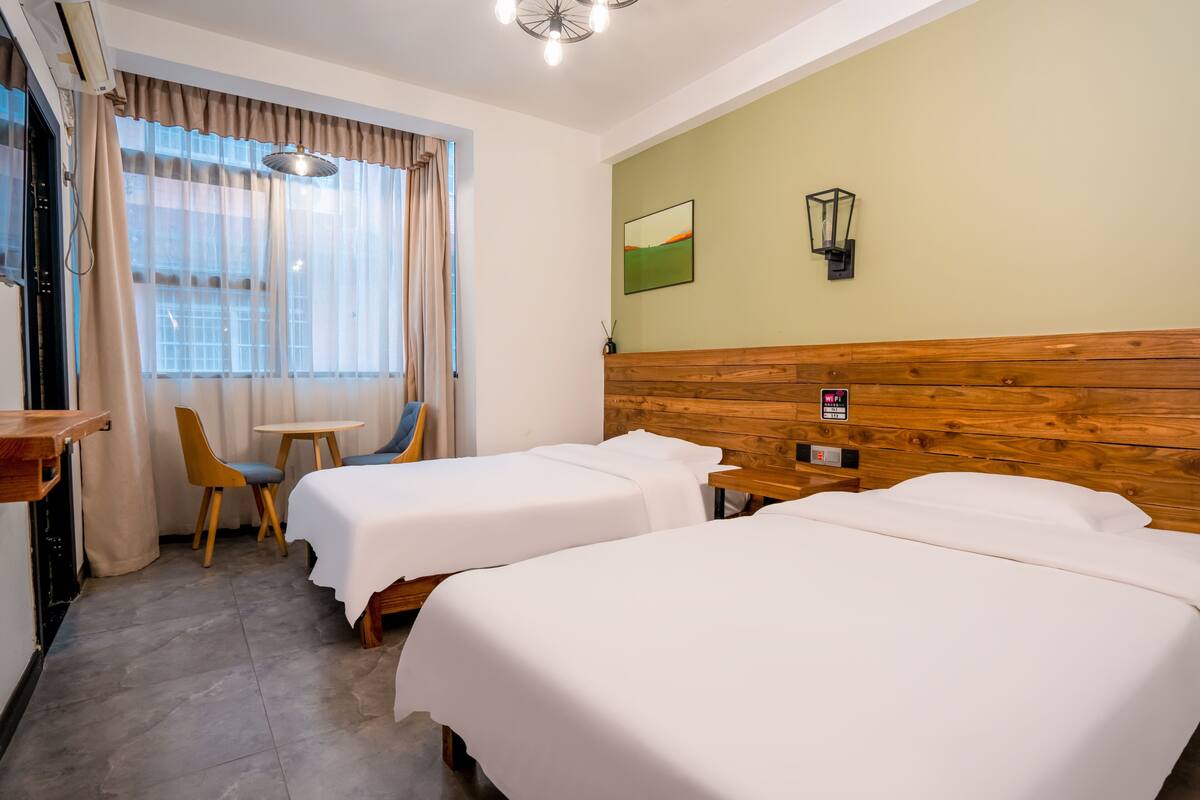 Ein Hotelzimmer mit zwei Betten, einem Holzkopfteil, einem kleinen Tisch, einem Stuhl und einem Fenster mit Vorhängen.