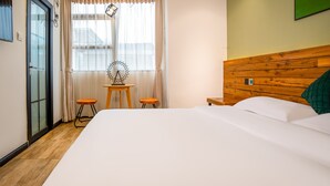 Habitación de hotel con una cama grande, cabecera de madera, una mesita con dos taburetes, una ventana con cortinas y una obra de arte colgada en la pared.