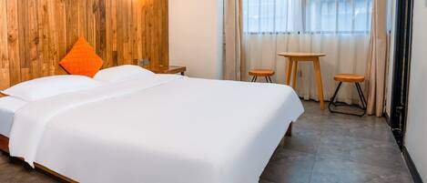A neatly made bed with white bedding, a wooden headboard, and an orange pillow. There are two small wooden stools and a table near a window with sheer curtains.