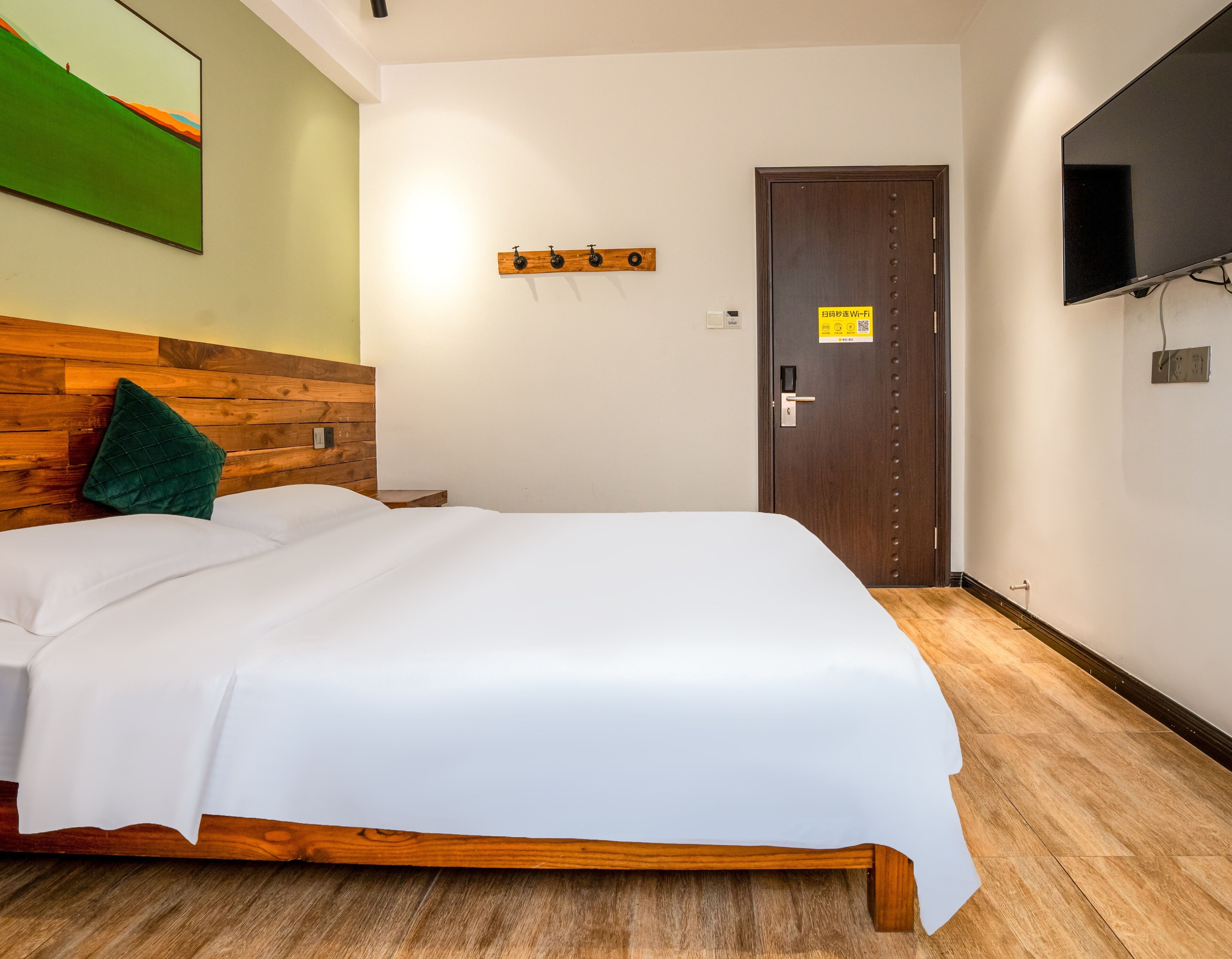 A neatly made bed with a wooden headboard, a green pillow, and a white bedspread. There is a wooden nightstand beside the bed, a wall-mounted TV, and a door with a sign indicating Wi-Fi availability.
