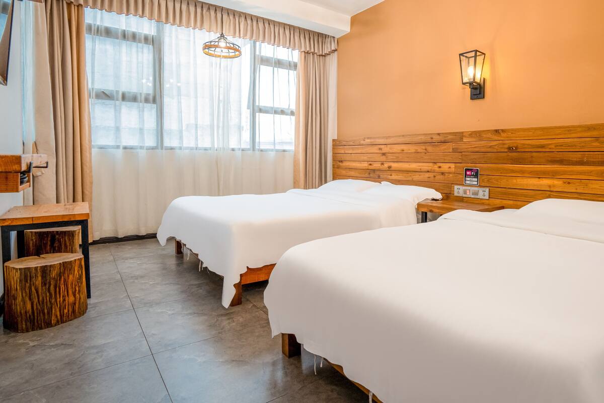 Ein Hotelzimmer mit zwei Betten, einem Holzkopfende, einem Nachttisch, einem kleinen Holzschemel und einer Wandlampe.