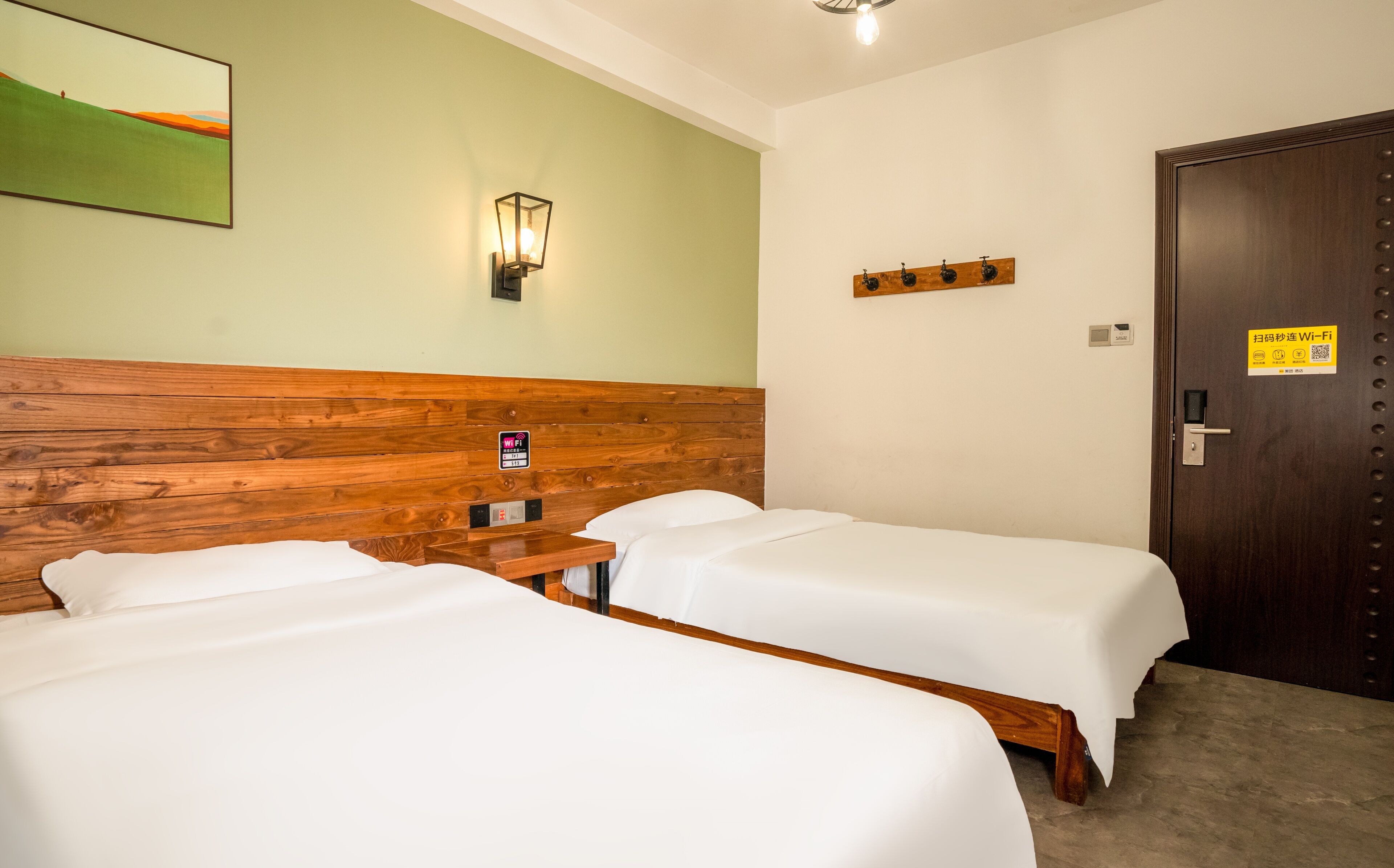 Quarto de hotel com duas camas de solteiro, cabeceira de madeira, luminária fixada na parede, porta com o sinal de Wi-Fi e uma mesinha.