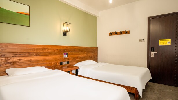 Une chambre d’hôtel avec deux lits simples, une tête de lit en bois, une lampe fixée au mur, une porte avec un panneau indiquant la présence du Wi-Fi et une petite table.