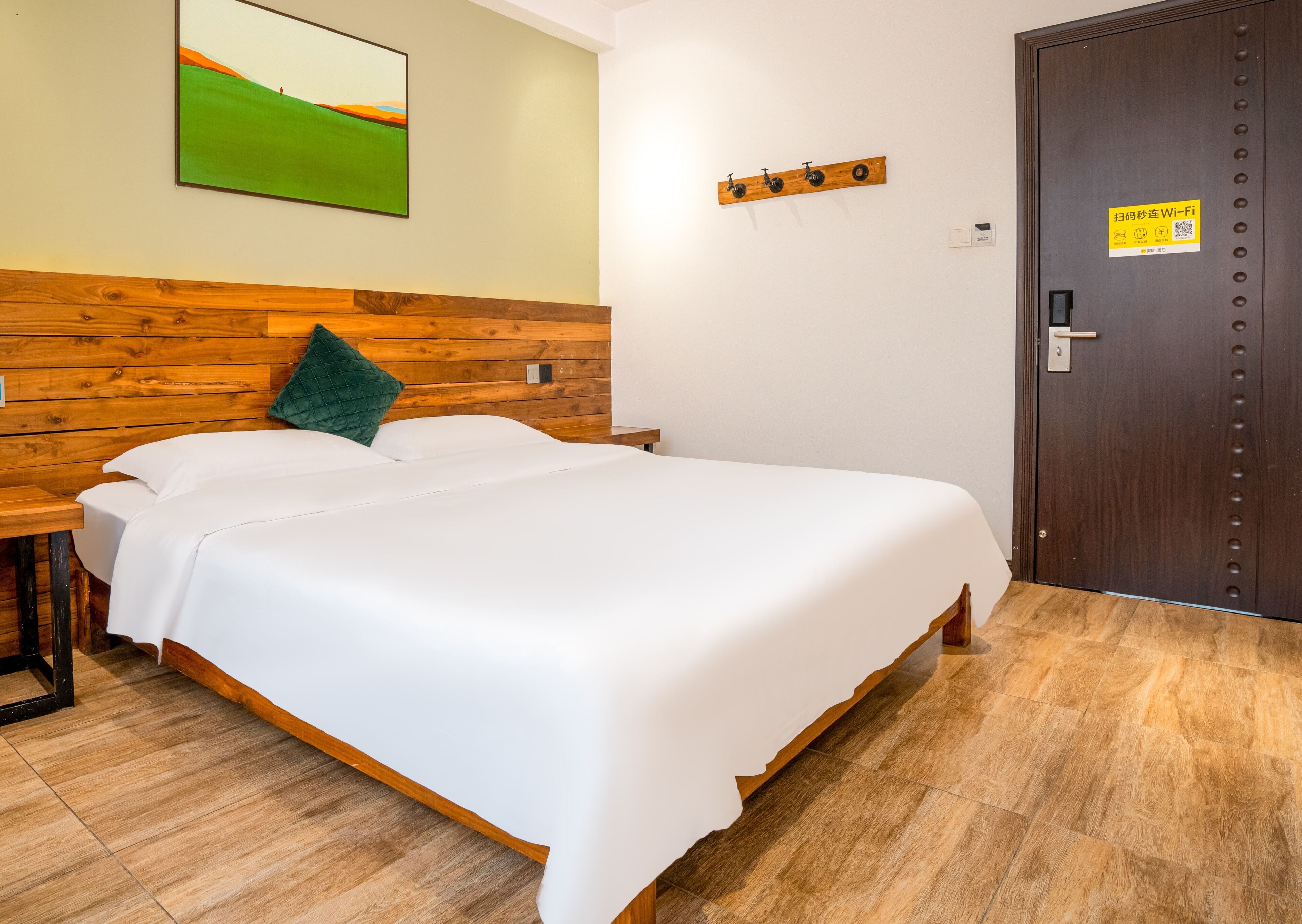 Ein Hotelzimmer mit Holz-Kopfteil, zwei Betten, einem grünen Kopfkissen, einem Bild an der Wand und einer Tür mit einem WLAN-Symbol.