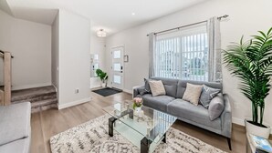 A modern living room with a grey sofa, a glass coffee table, and a large window with curtains. - New house to stay in Calgary close to Airport and the big mall Crossiron Mill (Calgary)