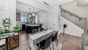 A modern kitchen with a dining area, featuring a long table, chairs, pendant lights, and a staircase. - New house to stay in Calgary close to Airport and the big mall Crossiron Mill (Calgary)