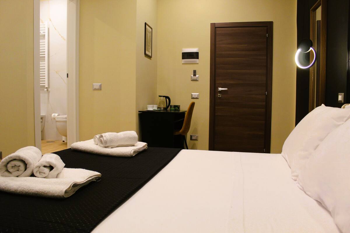 Ein Hotelzimmer mit einem Bett, einer schwarzen Matte, Handtüchern, einem kleinen Tisch mit einem Wasserkocher und einer Tür.