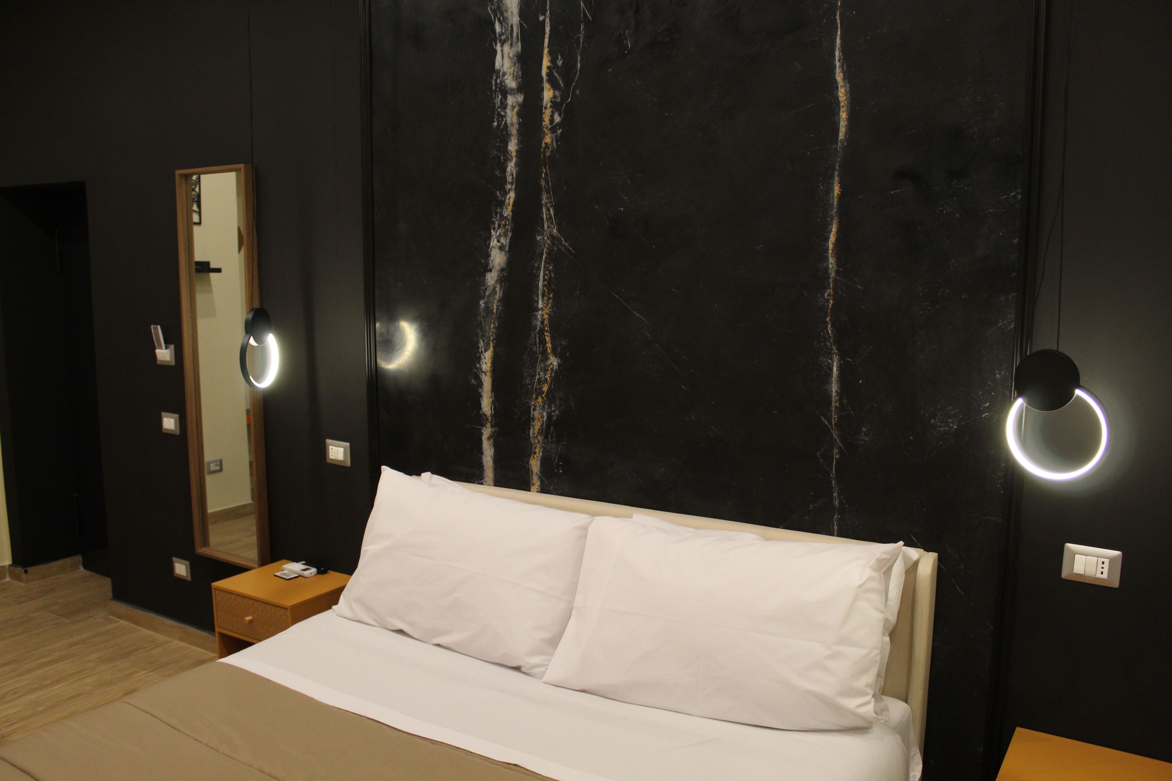 A hotel room with a bed, a nightstand, a mirror, and wall-mounted lighting.