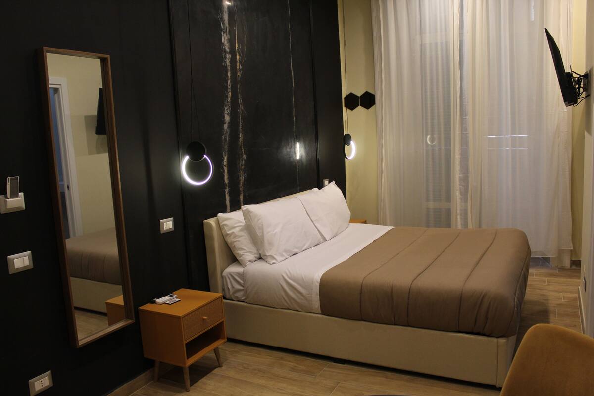Ein Schlafzimmer mit einem großen Bett, einem Nachttisch, einem Spiegel und einem Fenster mit Vorhängen.