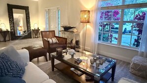 A living room with a sofa, a coffee table, a chair, a lamp, and a window with a view of flowers and a car. - House - Sleeps 6 - Hot Tub - Garden - Near Beach (Naples)