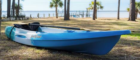 Un kayak azul con la marca 'feelfree' en un césped cerca de una masa de agua con palmeras y un muelle de madera al fondo.