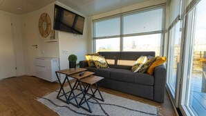 A living room with a sofa, a coffee table, a flat-screen TV mounted on the wall, and a large window with blinds.