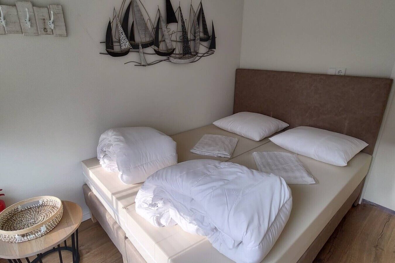 Una cama con ropa de cama blanca y cabecera, una obra de arte abstracto colgada en la pared, y una pequeña mesa redonda con una cesta tejida.