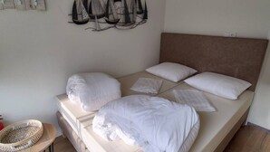 A bed with white bedding and a headboard, a wall-mounted abstract art piece, and a small round table with a woven basket.