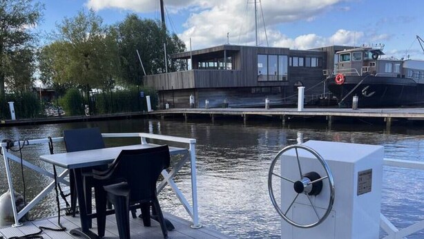 A wooden deck with a table and chairs, a white electrical box, and a boat docked in the background.